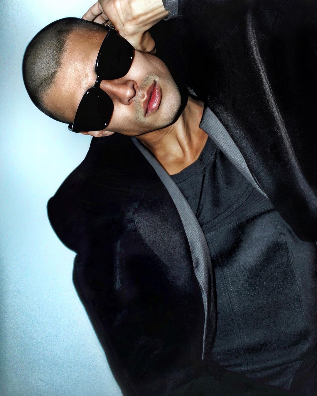 Close-up portrait of an adult man with short hair, wearing sunglasses and a dark jacket against a blurred studio background.