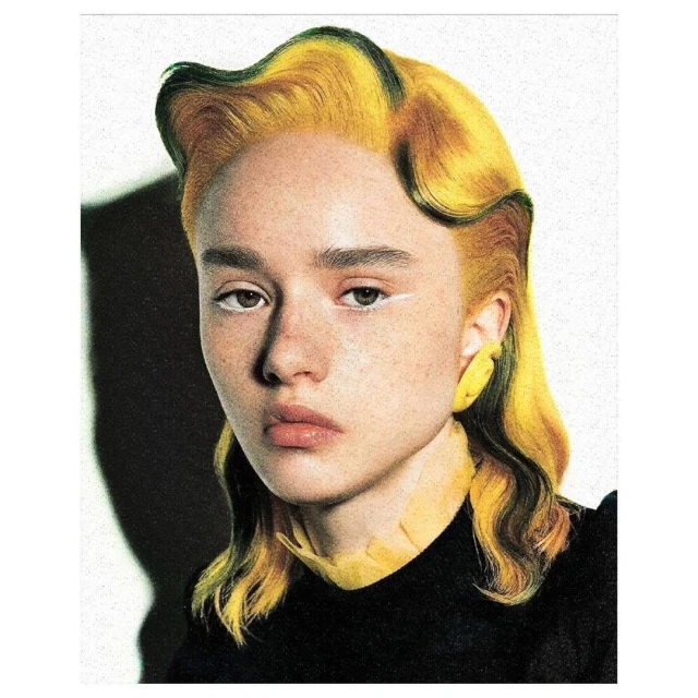 Portrait of a young woman with yellow wavy hair and white eyeliner, wearing a black top against a textured white background.