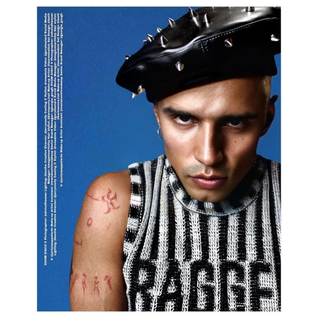 Close-up of a young adult male wearing a black studded beret and a striped shirt against a bright blue background.