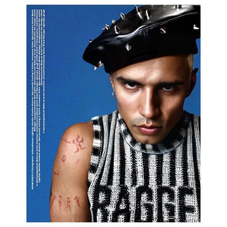 Close-up of a young adult male wearing a black studded beret and a striped shirt against a bright blue background.