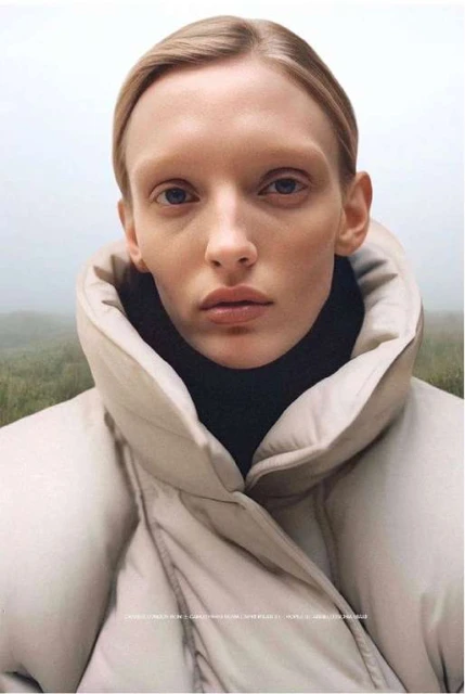 Close-up of a fair-skinned woman with light blonde hair in a beige puffer jacket against a misty, out-of-focus outdoor background.