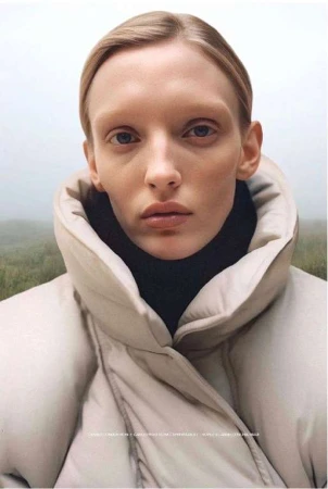 Close-up of a fair-skinned woman with light blonde hair in a beige puffer jacket against a misty, out-of-focus outdoor background.