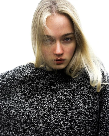 Close-up portrait of a woman with long blonde hair wearing a textured black and white sweater against a plain white background.