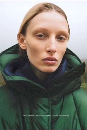 Close-up portrait of a blonde woman wearing a green hooded puffer jacket against an overcast misty outdoor background