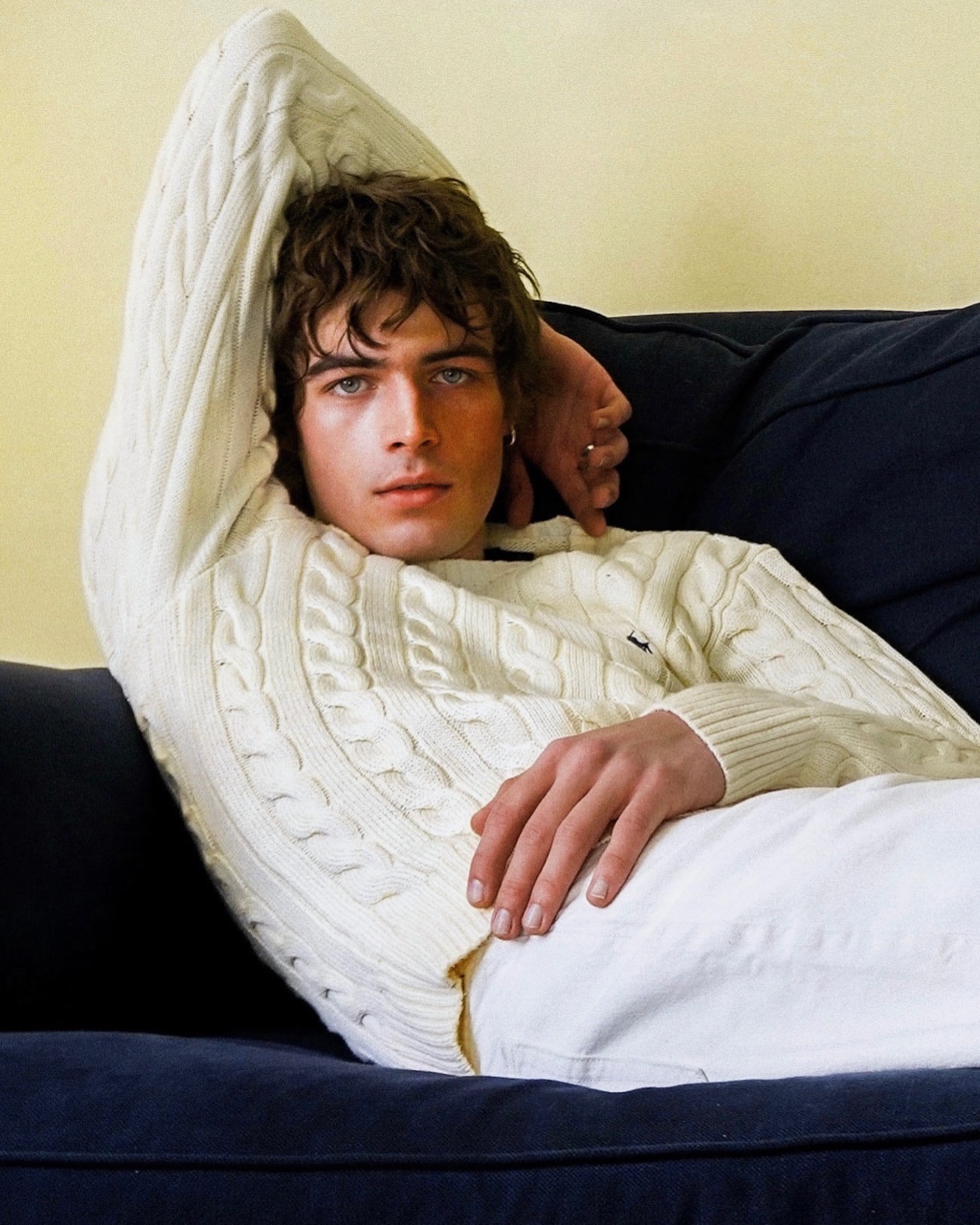 A young adult male model lies on a dark blue couch wearing a white cable knit sweater and white pants in a relaxed pose.