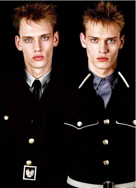 Close-up studio shot of two young adult men with fair skin and light hair wearing dark formal jackets with gold buttons against a black background.