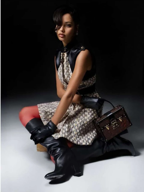 Portrait of a brunette woman sitting in a patterned dress and black leather vest with knee-high boots, holding a small box purse.
