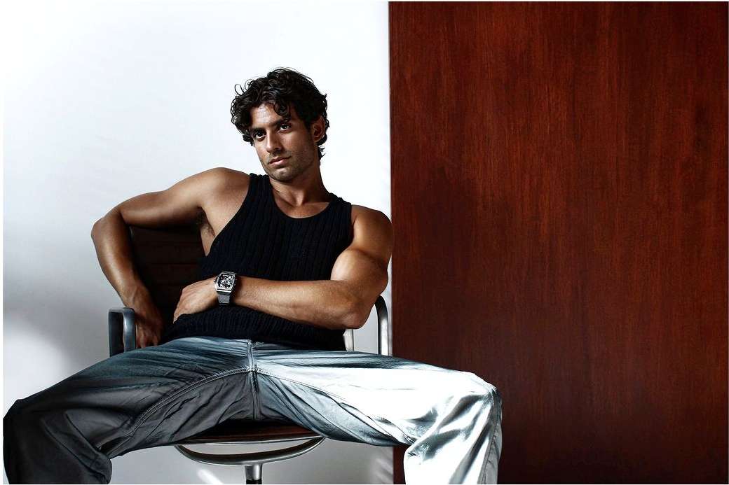 Portrait of a man with dark curly hair wearing a black tank top and silver watch seated in a chair by Ahmed Hassan