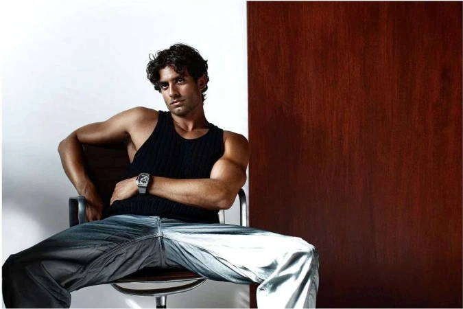 Portrait of a man with dark curly hair wearing a black tank top and silver watch seated in a chair by Ahmed Hassan
