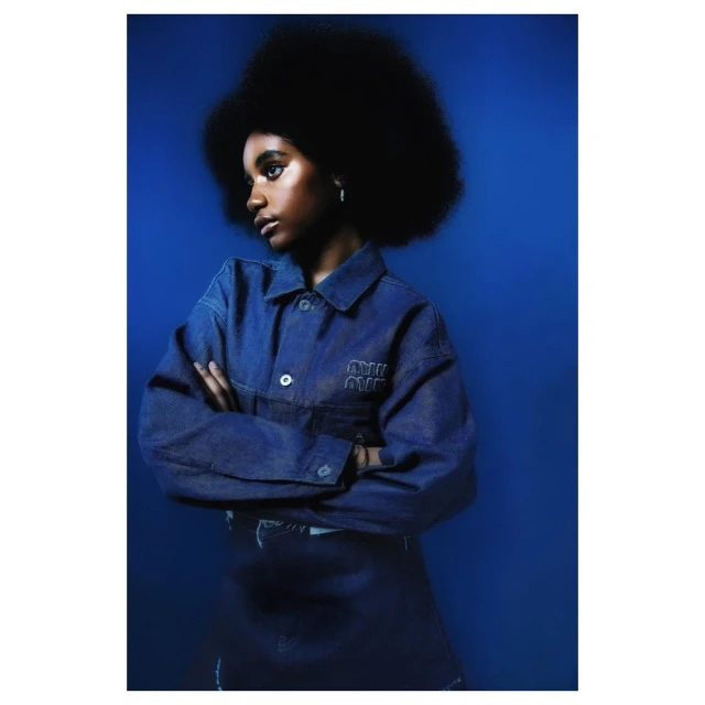 Teen girl with an afro poses in a studio wearing a denim jacket and skirt against a dark blue background in a medium studio shot.