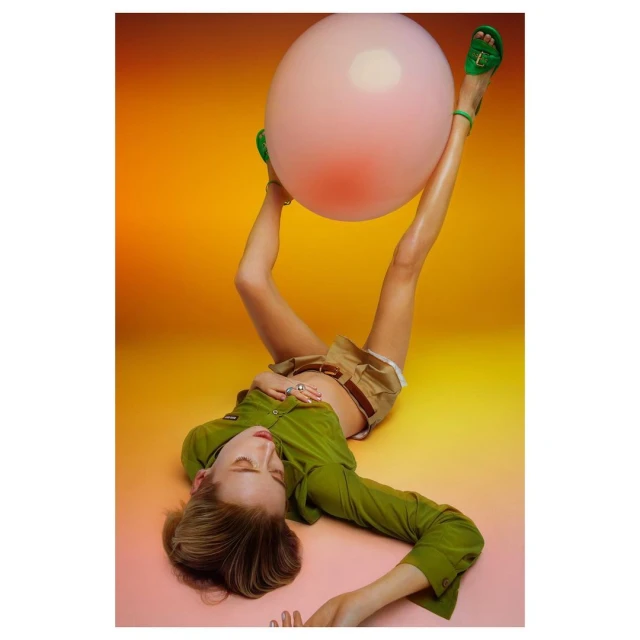 A blonde-haired woman in a green outfit and heels reclines with a large pink balloon against a gradient yellow background indoors.