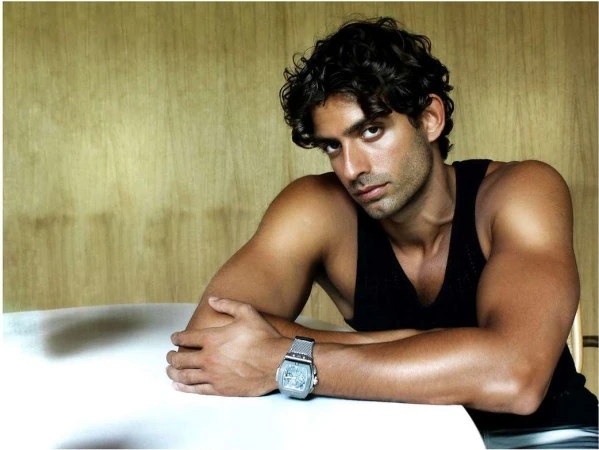 Man with dark curly hair wearing a black tank top and silver watch seated at a white table against a wood paneled wall.