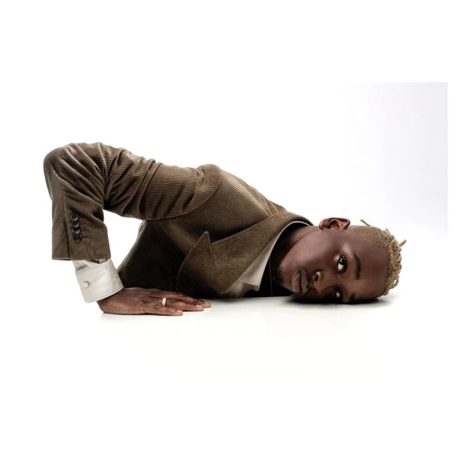 A young adult African American man lies on a white surface wearing a corduroy suit jacket, his head facing the camera in a studio setting.
