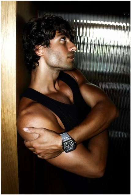 Attractive adult male with curly hair poses with crossed arms indoors wearing black tank top and wristwatch in studio lighting conditions.