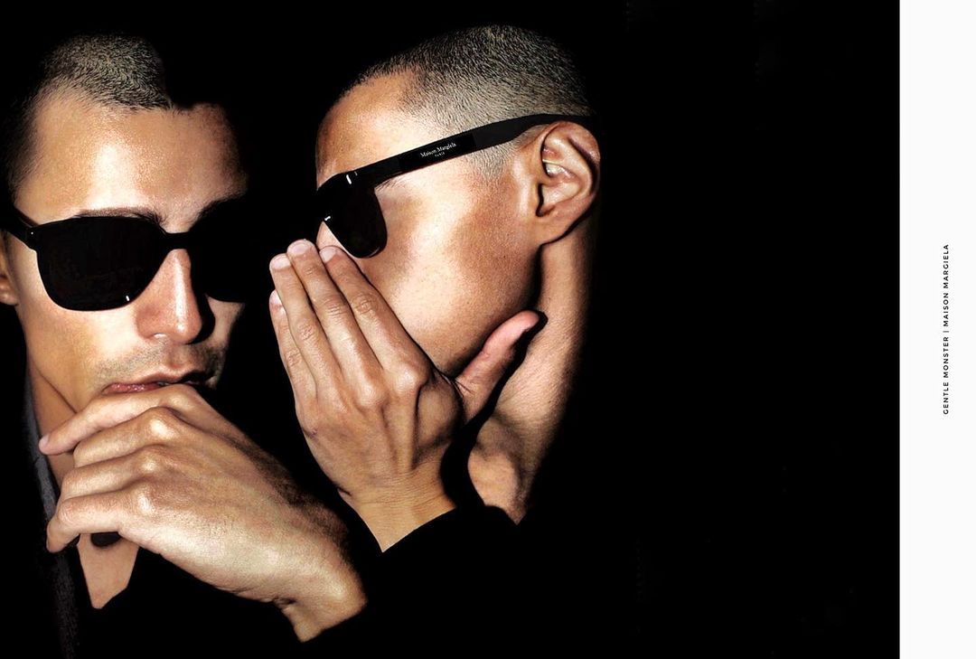 Two adult males wearing sunglasses pose indoors in front of a dark background, one with his hand to his mouth.
