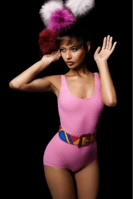 Fashionable woman in pink swimsuit and colorful belt strikes a pose against a black backdrop, adorned with a large, fluffy, whimsical headpiece.