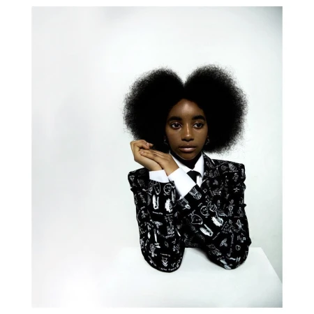 Portrait of a Black woman with voluminous afro hair wearing a black and white patterned blazer over a white shirt, posed against white seamless
