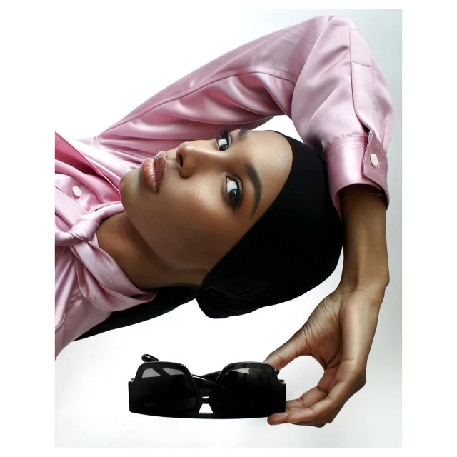 Elegant studio portrait of a woman with a black headscarf, wearing a pink satin top, holding black sunglasses against a white backdrop.