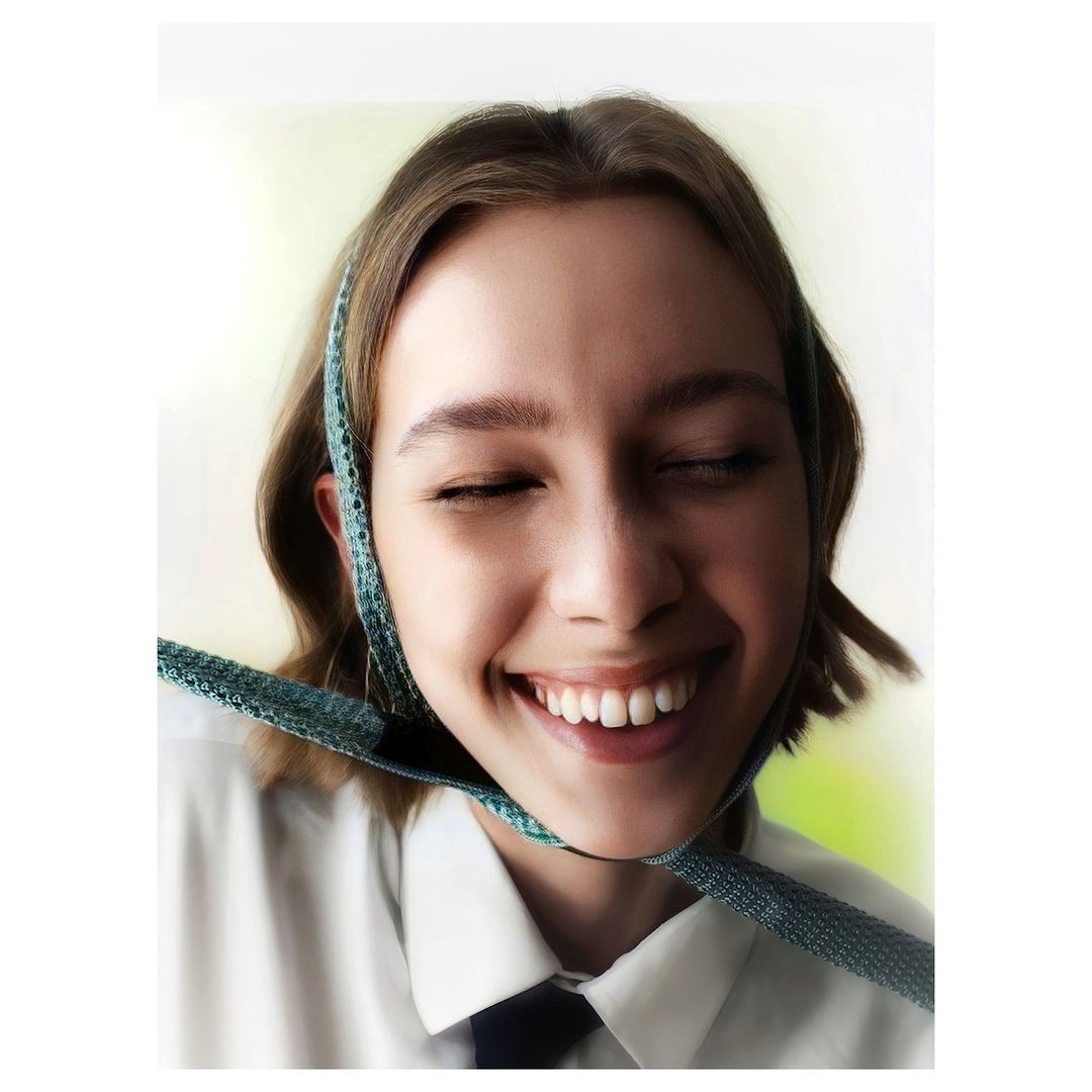 Close-up portrait of a smiling young woman with fair skin and brown hair, wearing a white collared shirt and dark necktie, eyes closed.