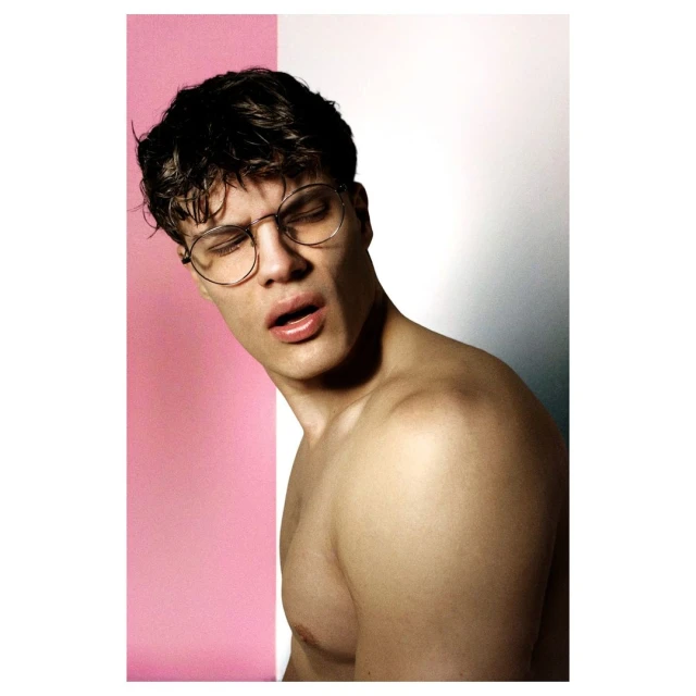 Portrait of a young adult man with dark hair and glasses against a split pink and white background, showcasing a fashion-forward aesthetic.