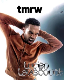 Man wearing a brown zip-up jacket posing with hands behind his head against a bright gradient background.