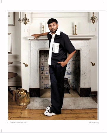 Man wearing a black and white shirt and dark trousers leaning on a marble fireplace mantel inside a room