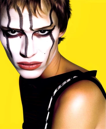 Close-up portrait of a model with dramatic face paint and dark eyeliner against a solid vibrant yellow background.
