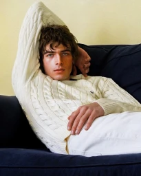 Young man in a white cable-knit sweater reclining on a dark blue sofa against a pale yellow wall