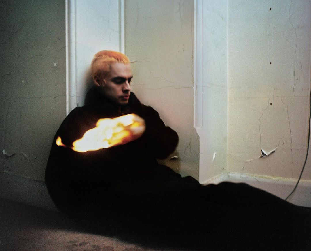 Young adult male with dyed hair poses against a white wall indoors, wearing a black jacket with an orange light effect. Moody setting.