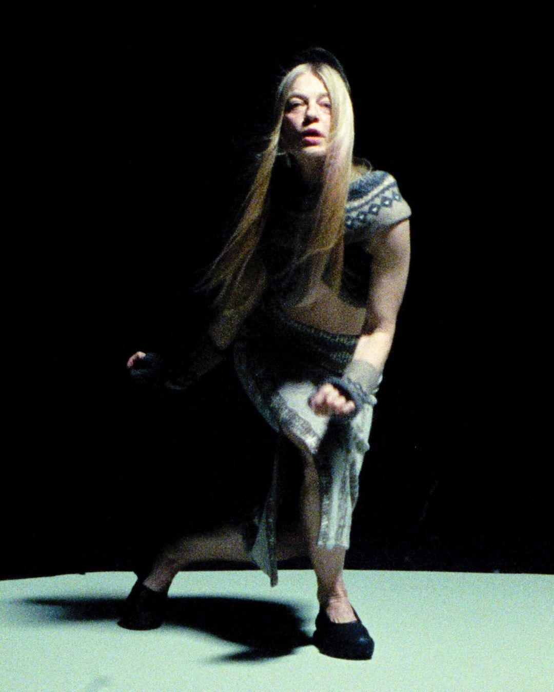 An adult female with long blond hair and a sweater poses in a crouching position in a photo studio with a black backdrop and low-key lighting.