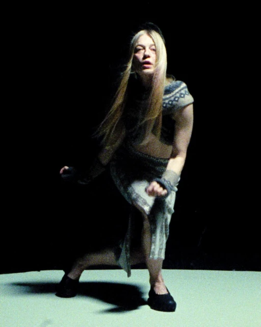 An adult female with long blond hair and a sweater poses in a crouching position in a photo studio with a black backdrop and low-key lighting.