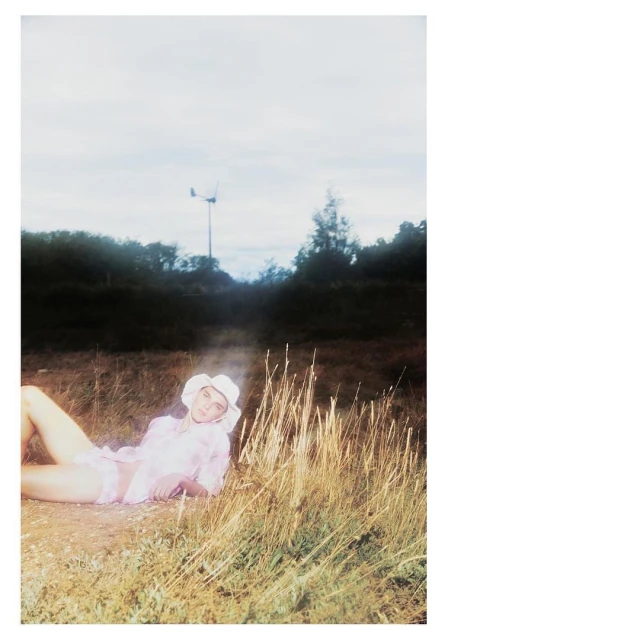 Woman in a white sun hat and pink clothing lies on a field of tall grass in front of a dark tree line with a bright sky.