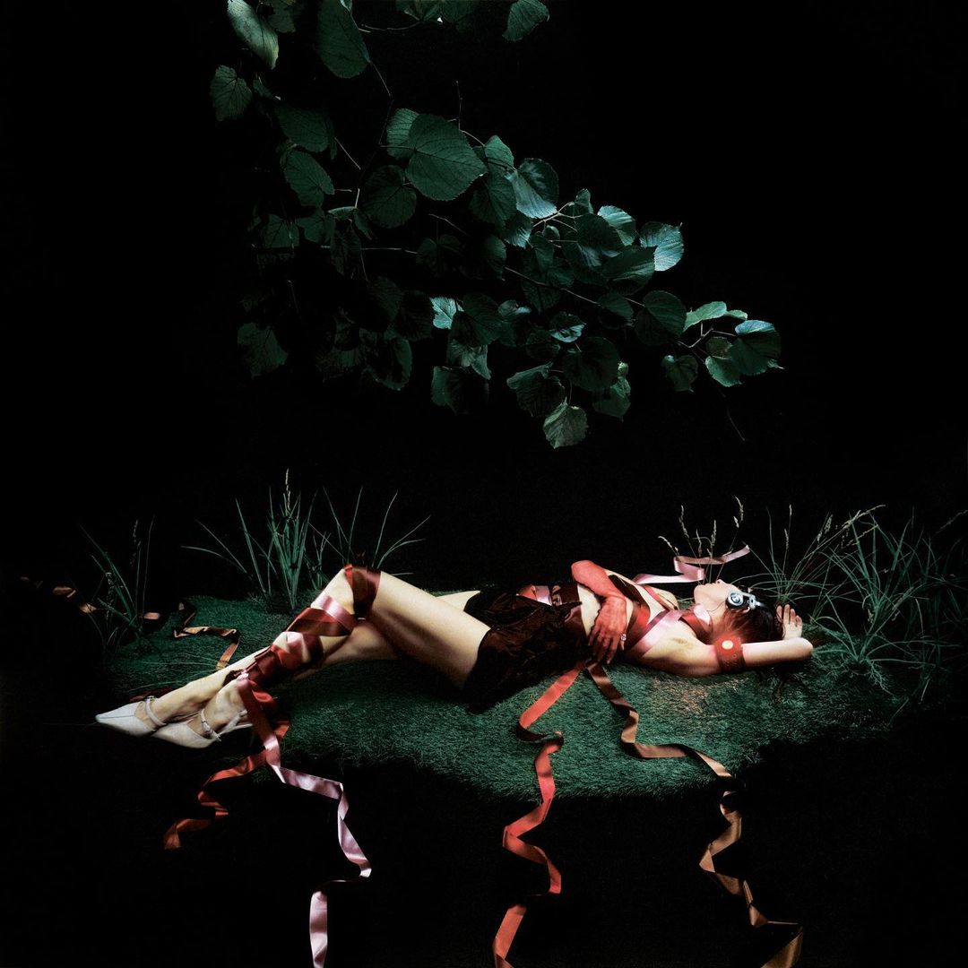 Woman lies on artificial turf surrounded by ribbons in a dark studio, lit with green foliage and studio lighting. Dark and fantasy aesthetic.