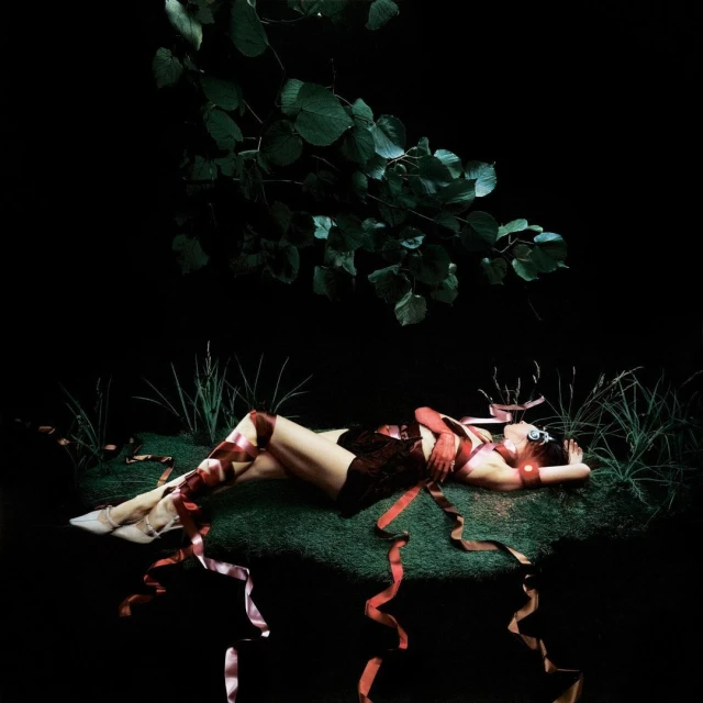 Woman lies on artificial turf surrounded by ribbons in a dark studio, lit with green foliage and studio lighting. Dark and fantasy aesthetic.