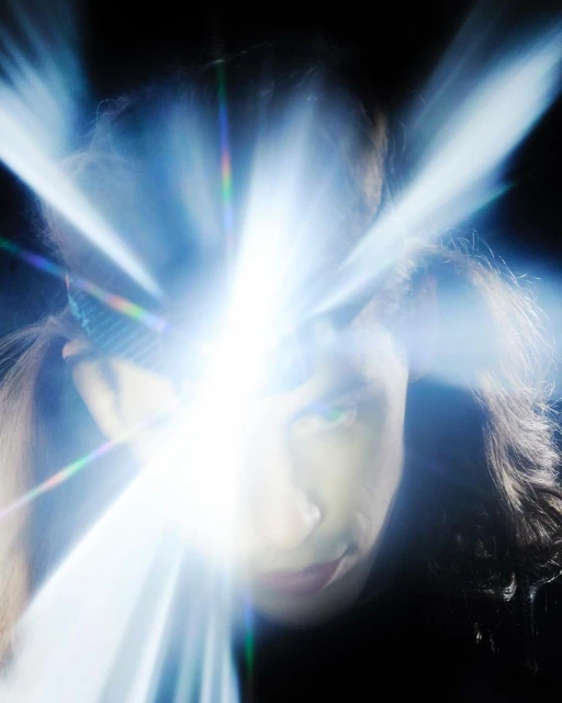 Close-up portrait of an adult with long hair and a headband, bathed in bright, directed light against a stark black background.