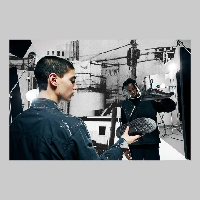 Two adult men holding black sneakers in a photography studio, with black and white city backdrop and studio lighting equipment.