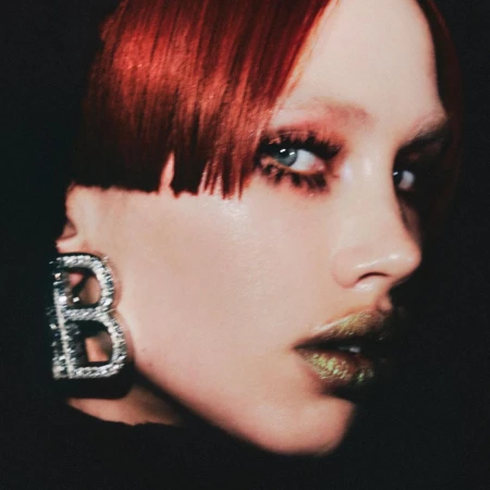 Close-up portrait of a woman with vibrant red blunt-cut hair wearing a large jewelled letter B earring.