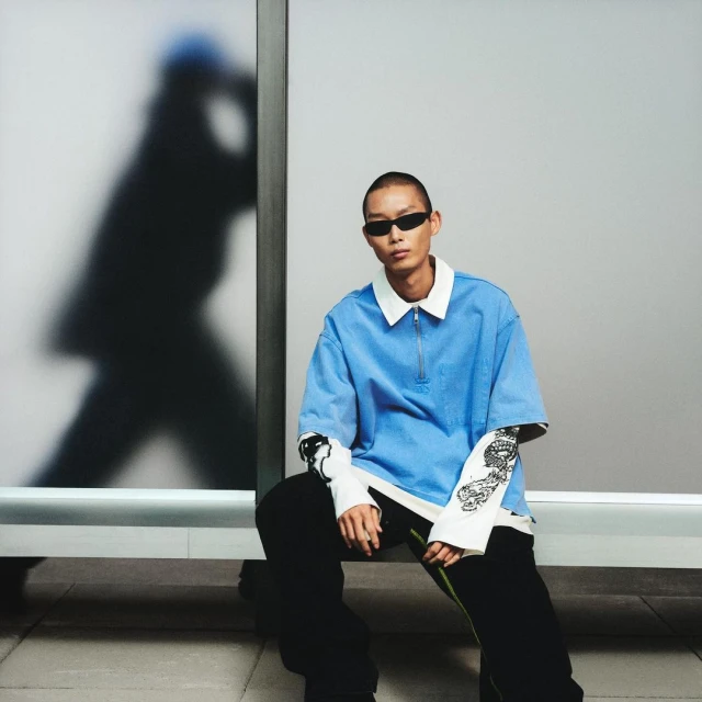 Stylish adult male seated, showcasing a light blue short-sleeve zip polo and black trousers, complemented by a graphic long-sleeve undershirt.