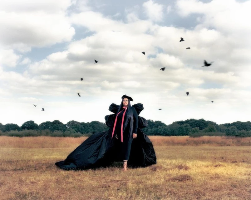 Fashionable woman in dramatic black coat with ribbons standing in field with birds flying above against cloudy sky, outdoor editorial.