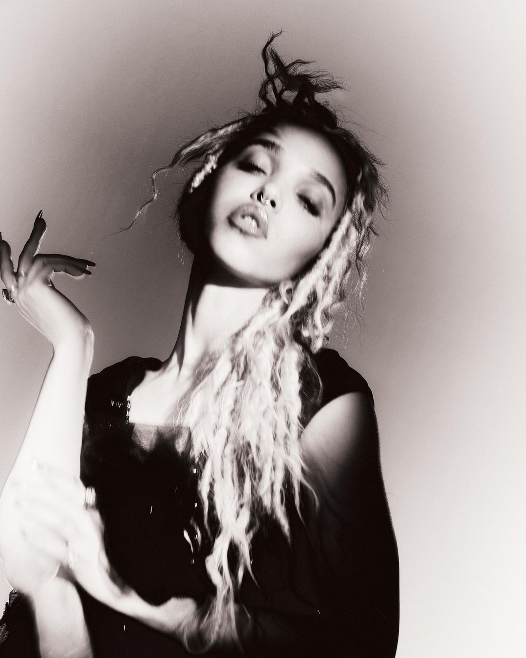 Monochromatic portrait of a young adult female with dreadlocks, facing the camera with a slightly pouting expression in a dimly lit setting.