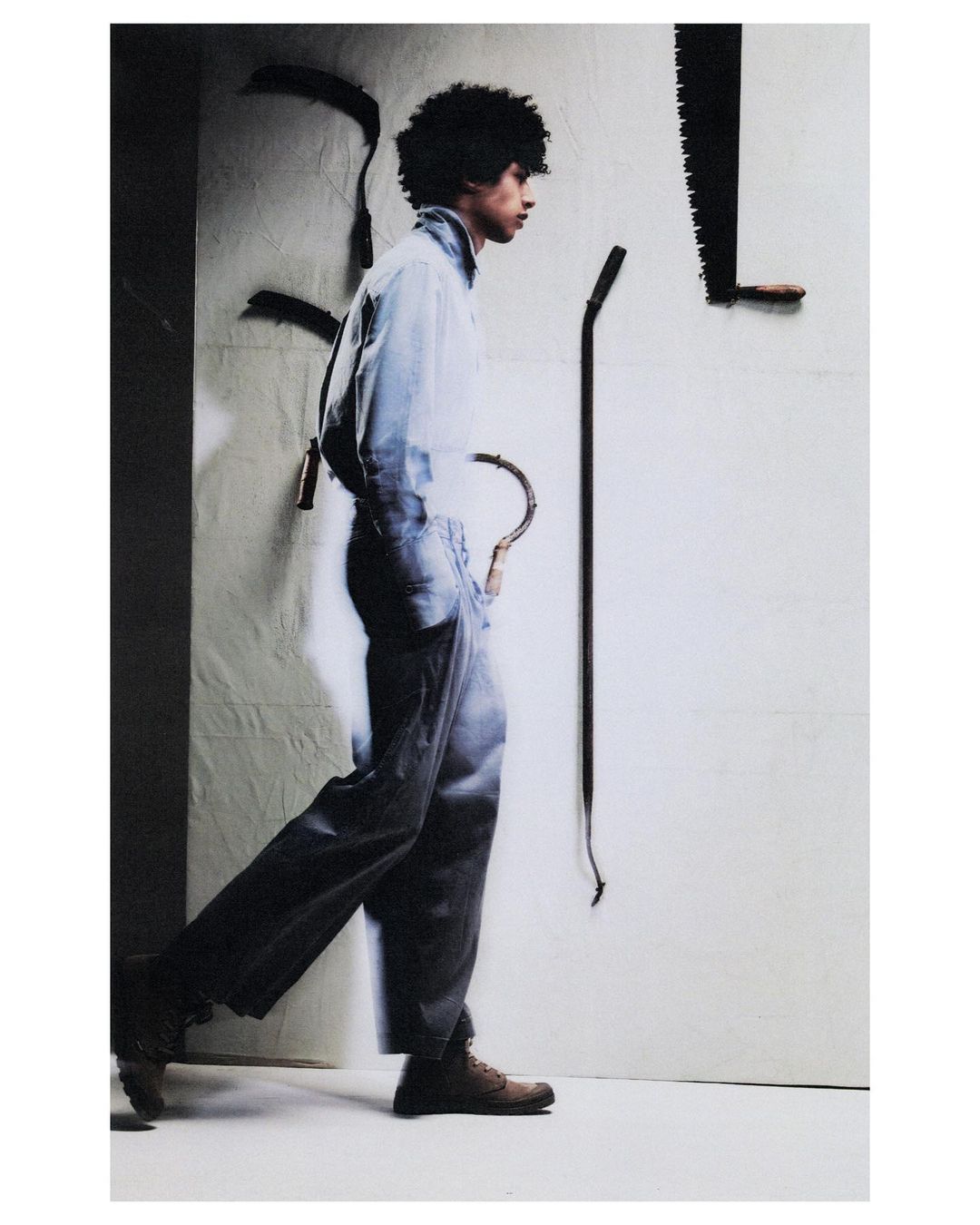 Adult male with afro walks in blue workwear. Displayed against white wall with mounted antique farming tools. Studio lighting.