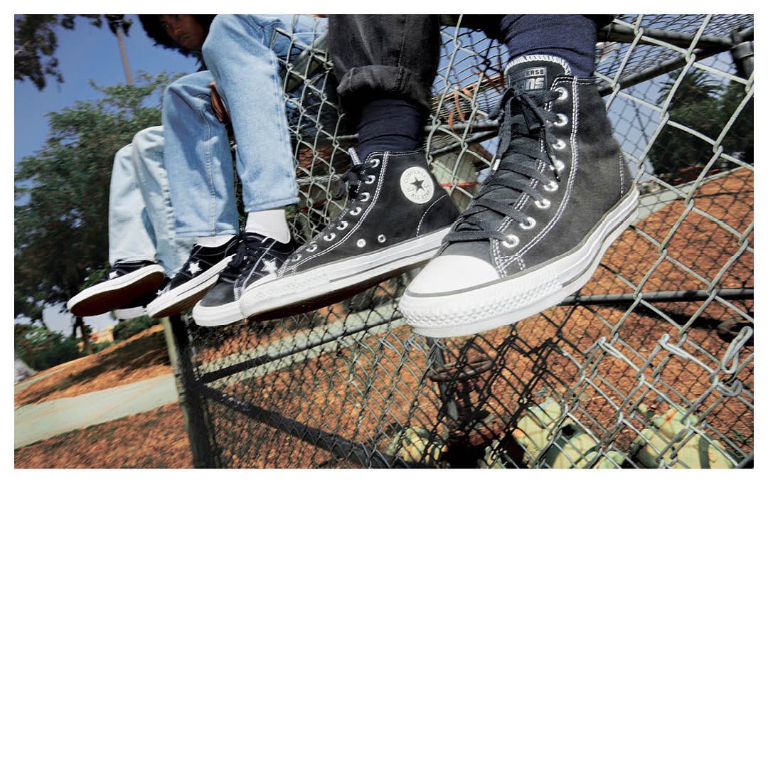 Three pairs of black and white Converse sneakers on a metal fence, showcasing high-top and low-top styles with blue skies and trees in the background.