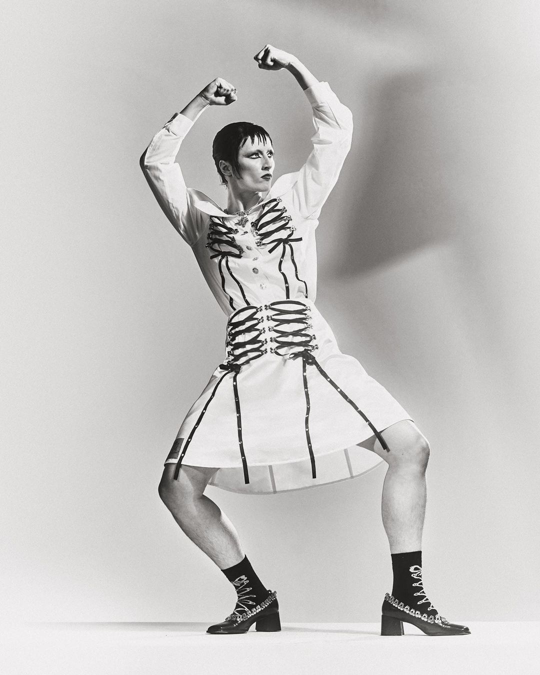 Person in white dress with black skeleton design, black socks and heels, posing against a white background in a studio.