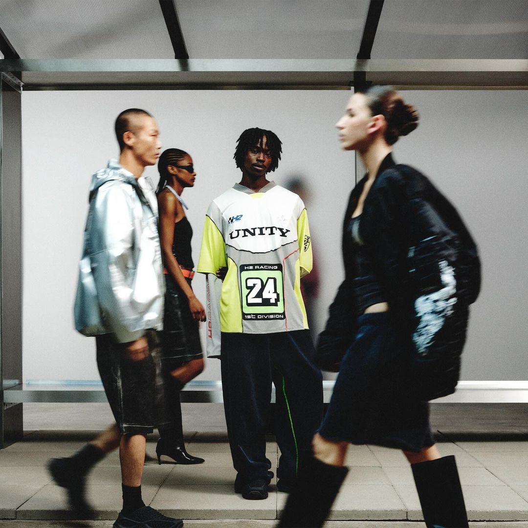 Four people wearing contemporary fashion stand near a bright wall. One person in the foreground walks to the right. Fashion shot.