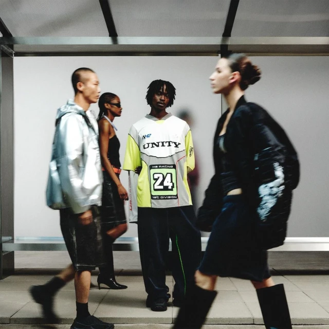 Four people wearing contemporary fashion stand near a bright wall. One person in the foreground walks to the right. Fashion shot.