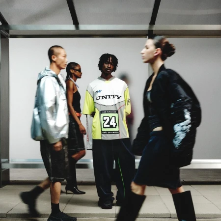 Four people wearing contemporary fashion stand near a bright wall. One person in the foreground walks to the right. Fashion shot.