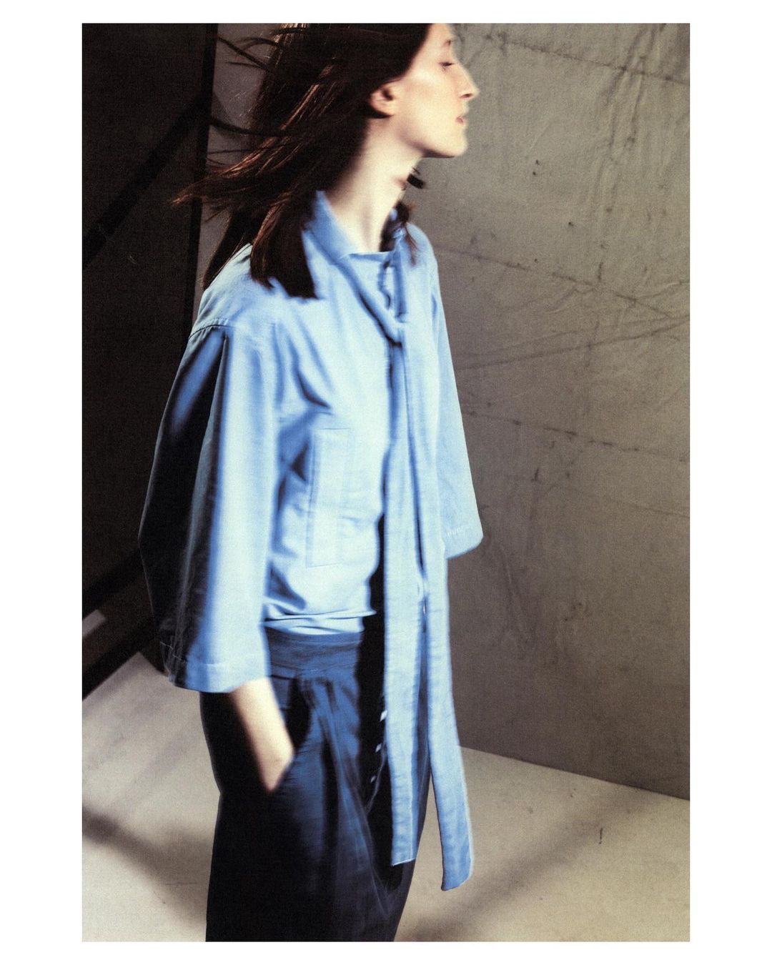 A slender woman in a light blue top and dark blue pants stands in a studio setting with a textured gray backdrop, casual and stylish.