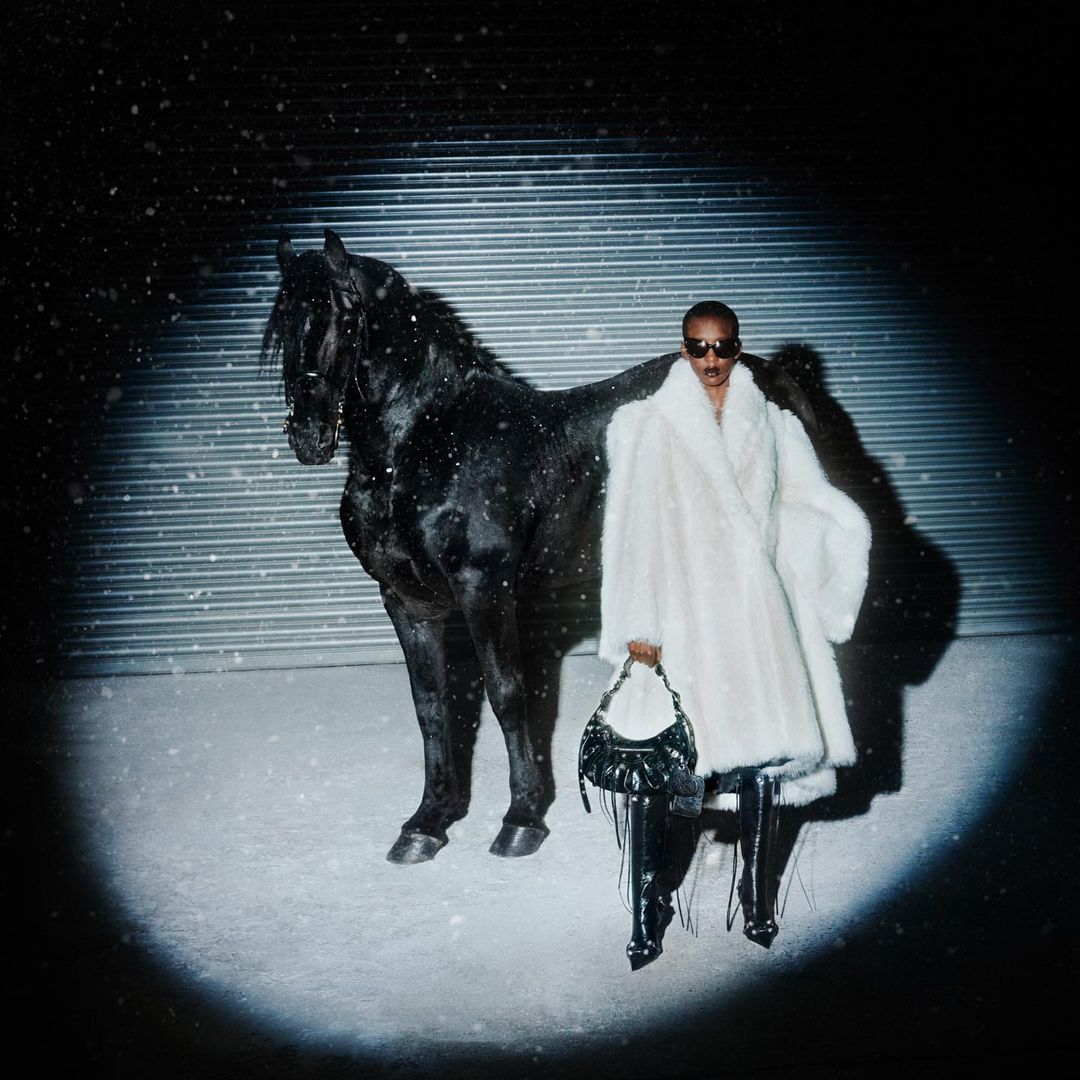 A person wearing a white fur coat poses next to a black horse against a metallic background with falling snow in a nighttime scene.