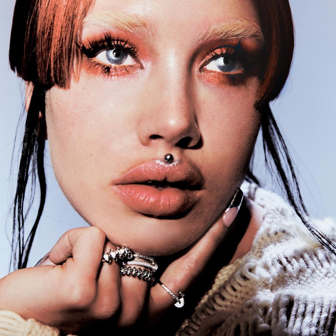 Close-up portrait of a woman with blue eyes, red hair, lip piercing, wearing multiple rings and a knitted sweater against a light blue background.