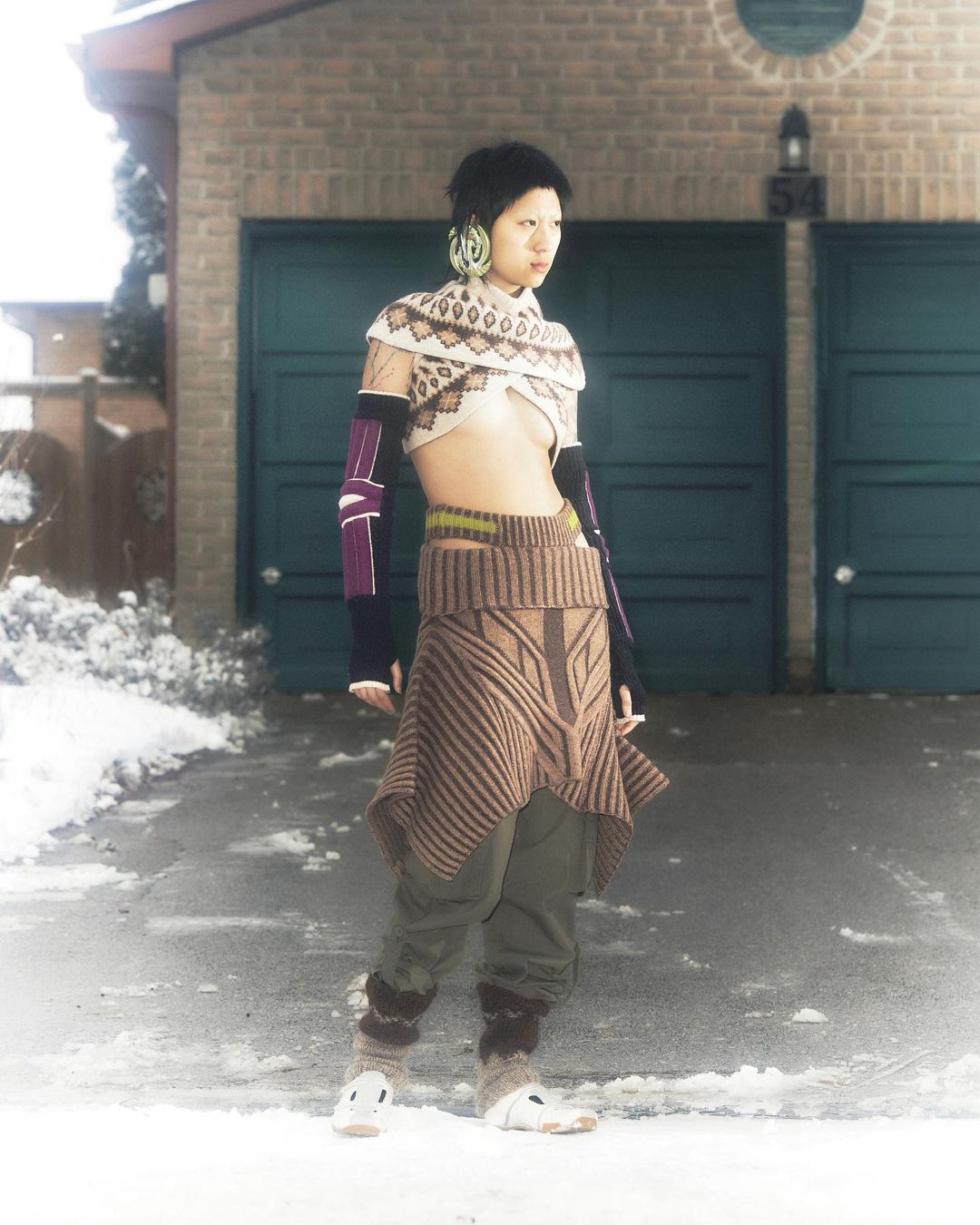 Asian woman in knitwear fashion poses in front of a garage in winter. She's wearing a crop top, skirt, arm warmers, and leg warmers.